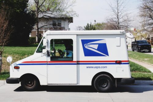 USPS mail truck with blue logo and red stripe parked on a suburban street. Trees and houses in the background under a clear sky.
