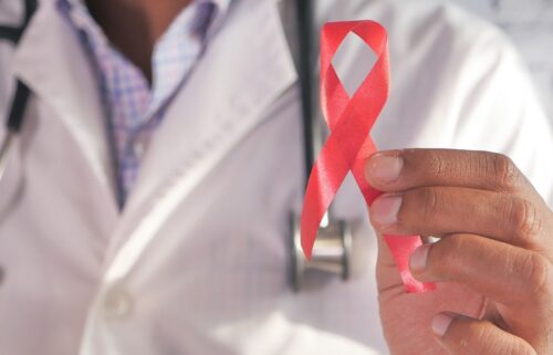 A person in a doctor's coat holds a red awareness ribbon between their fingers, symbolizing support and awareness for HIV/AIDS. The tone is supportive.