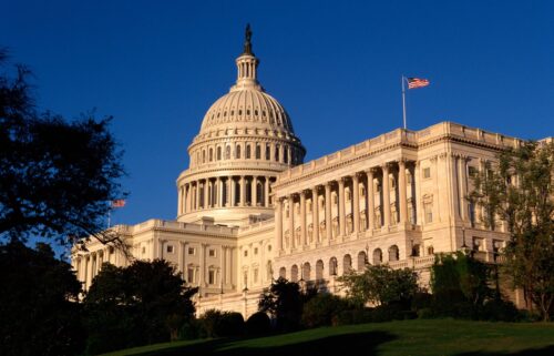 The U.S. Capitol building under a clear blue sky, featuring its iconic dome and columns with an American flag waving, surrounded by lush greenery.