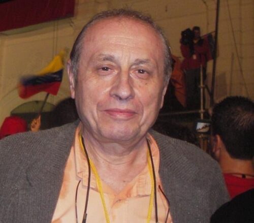 A middle-aged man with graying hair stands amidst a crowd. He is wearing a gray sports jacket and a peach button-down shirt. His glasses hang below his neck, out of sight.
