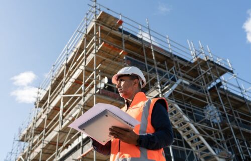 A construction worker in a hard hat and orange vest holds a tablet while examining a scaffolding structure under a clear blue sky, representing just one of the millions in low-benefit industries like restaurants and construction may be left uninsured and without Medicaid.