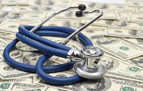 A blue stethoscope rests atop a scattered pile of U.S. hundred-dollar bills, symbolizing the financial aspects of healthcare and the damage done if medicare is cut. The tone is serious and thought-provoking.