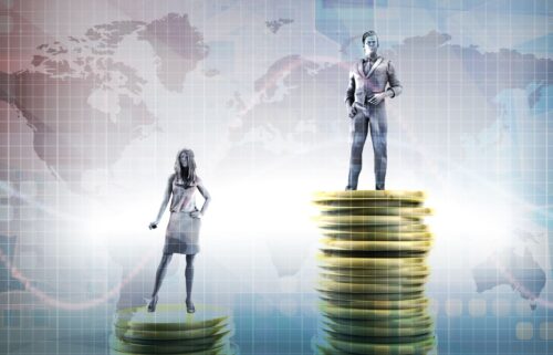 A woman and a man stand on separate stacks of coins against a world map backdrop. The man stands on a taller stack, suggesting a budget or pay gap theme.