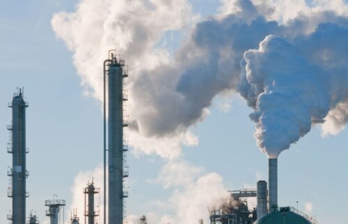 Industrial chimneys releasing thick white smoke against a clear blue sky, highlighting pollution and environmental impact. The scene conveys industrial activity.