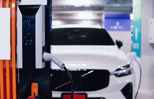 Electric vehicle charging at a station in a parking garage. The white car is connected to a charger with a visible charging plug, conveying a modern, eco-friendly tone.
