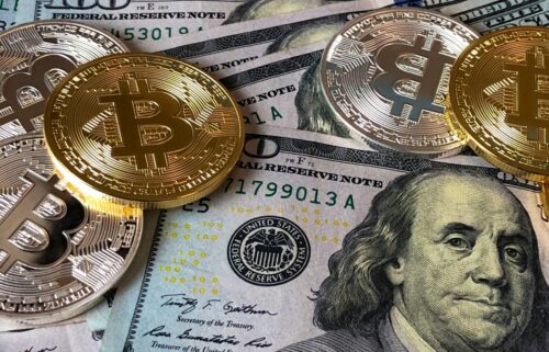 Gold and silver Bitcoin coins rest on U.S. hundred-dollar bills, featuring Benjamin Franklin. The image contrasts traditional and digital currencies.