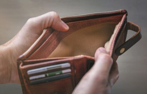 Hands holding an open, empty brown leather wallet, highlighting financial emptiness. Three cards visible in the slots, with an overall tone of scarcity and concern.