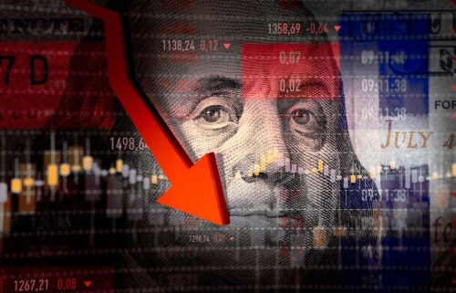 A collage of a downward red arrow overlaying Benjamin Franklin's face on a hundred-dollar bill, with stock market graphs and numbers in the background, depicting financial spending decline.