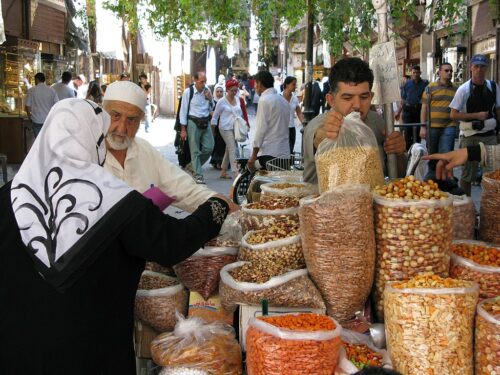 Street market scene with clear plastic bags of grains, nuts, and dried fruit appearing in the foreground. A man in a white shirt and white hat with a white beard talks to a woman in black whose back is turned toward the camera, while another man with dark hair in a gray shirt grabs a bag of grain. Dozens of shoppers and pedestrians appear in the background.