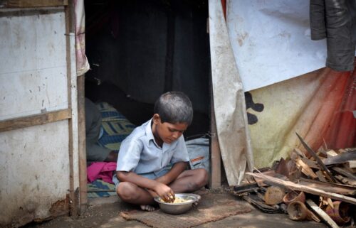 A young boy sits cross-legged outside a makeshift shelter, focused on eating from a metal bowl. Surrounding him are wooden planks and a patched curtain doorway in a country left in poverty due to debt.