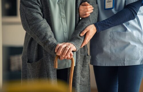 Elderly person with a cane is gently supported by a home health care worker, both wearing cardigans.