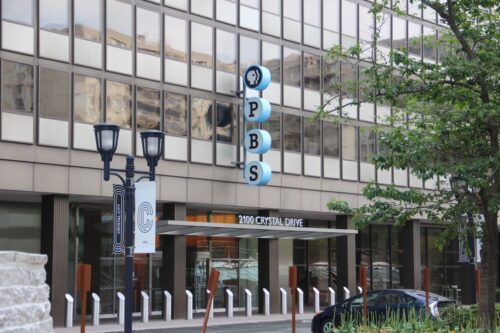 A Modern building exterior with large windows and a vertical PBS sign. (Public Broadcasting System) A street lamp and tree are in the foreground. The address reads 