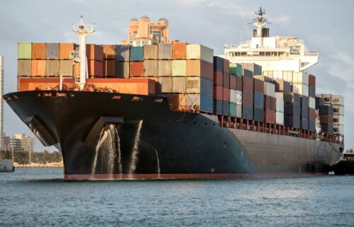 Large cargo ship carrying colorful containers sails through calm water, with an urban skyline in the background. GDP