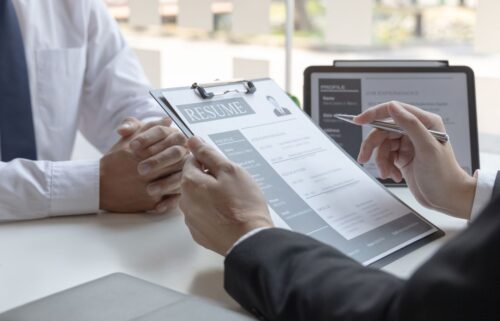 A businessperson in a suit reviews a resume during a a job interview. Hands folded on the table, showcasing a professional setting. Tablet with profile in background.