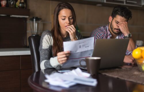 A worried couple sits at a kitchen table cluttered with bills and a laptop. The woman reads a document intently, while the man rubs his forehead, appearing stressed.