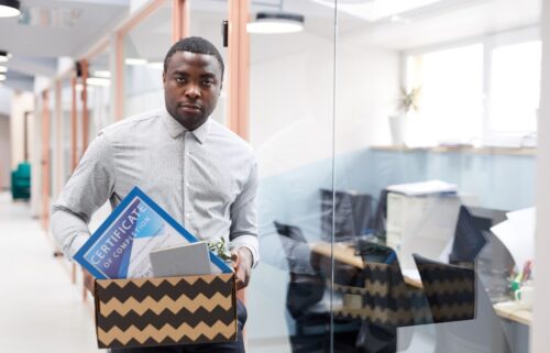 A man in a formal shirt holds a chevron-patterned box with a certificate, a plant, and other office belongings standing near an office's glass wall as he exits his job.