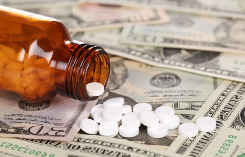 An amber pill bottle spills white tablets onto various US dollar bills, symbolizing the financial cost of healthcare or medication.