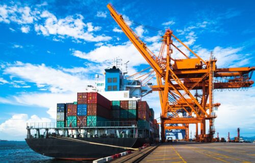 Cargo ship docked at a port under a clear blue sky, with colorful stacked containers and large orange cranes, conveying a sense of industrious activity.