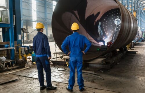 Two workers in blue uniforms and yellow helmets stand in a manufacturing factory, observing a large metal cylinder with a welder working inside, emitting sparks.
