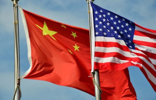 Two flags side by side against a blue sky: the red flag of China with yellow stars, and the United States flag with stars and stripes, symbolizing international relations.