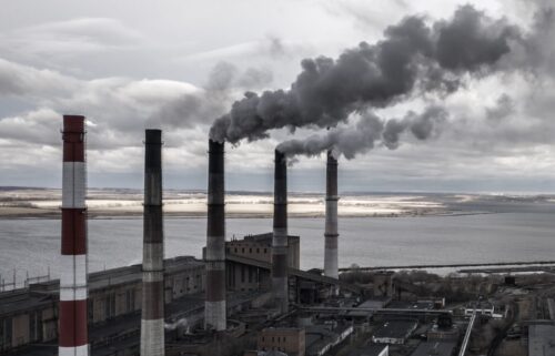 Industrial plant by a large body of water on a cloudy day. Five smokestacks emit thick, dark smoke, creating a gloomy, polluted atmosphere.