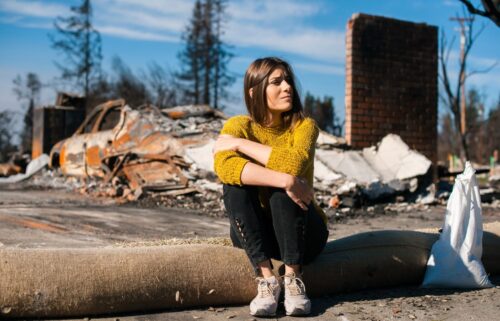 A woman in a yellow sweater sits pensively on a rolled-up carpet amidst debris from a fire waiting for FEMA Aid. Burnt car and collapsed structures in the background convey desolation.