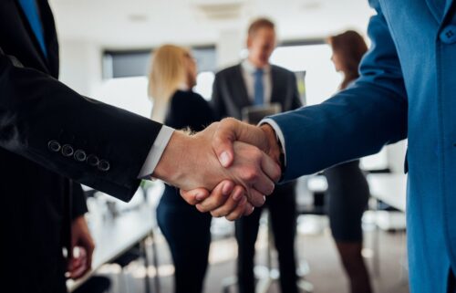 Two people in business attire shaking hands, suggesting how big pharma bought the government. Three other colleagues converse in the blurred background.