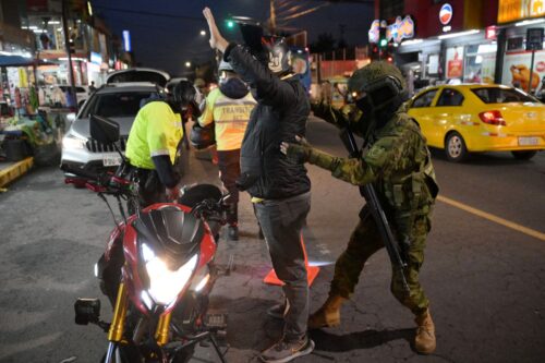 An Ecuadorian soldier in camouflage and helmet searches a person with raised arms at night, near a red motorcycle as new laws from President Noboa take effect. Nearby, traffic officers check a car on a busy street. (Photo by Rodrigo BUENDIA / AFP) (Photo by RODRIGO BUENDIA/AFP via Getty Images)