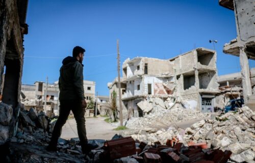 A man in a dark jacket stands among rubble in a war-torn city. Damaged buildings and debris surround him under a clear blue sky, evoking devastation.