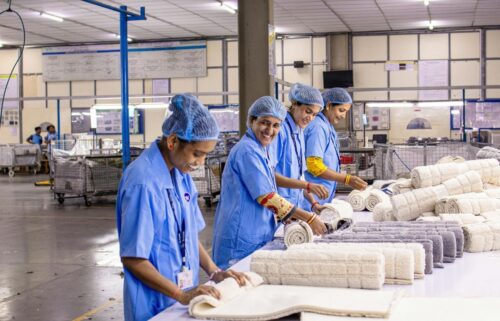 Four women in blue uniforms and hairnets smiling while rolling towels in a factory. The atmosphere is industrious and positive, with neatly organized stacks.