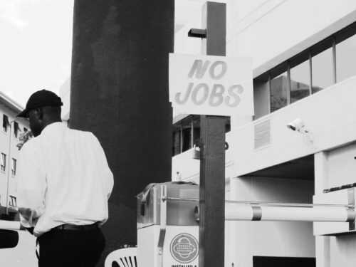 Black and white photo of a Black man in a white shirt and cap walking past a sign saying 