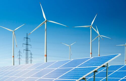 Solar panels and wind turbines under a clear blue sky, conveying a clean energy theme. Power lines in the background add to the sustainable energy context.