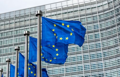 European Union flags, blue with yellow stars, wave in front of a modern glass building. The scene conveys a sense of unity and formality.
