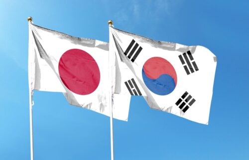 Two flags wave against a clear blue sky: the Japanese flag with a red circle on a white field, and the South Korean flag with a red and blue yin-yang and black trigrams.