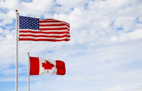 Two national flags on poles against a cloudy sky. The US flag waves above, featuring stars and stripes; below, the Canadian flag with a red maple leaf.