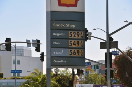 Gas station sign displaying prices: Regular $5.29, Plus $5.49, V-Power $5.69. Urban setting with palm trees and street signs in the background.