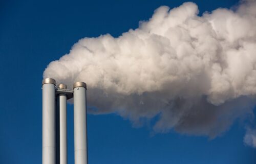 Three smokestacks emit thick white smoke emissions against a clear blue sky, symbolizing industrial pollution and environmental concerns.