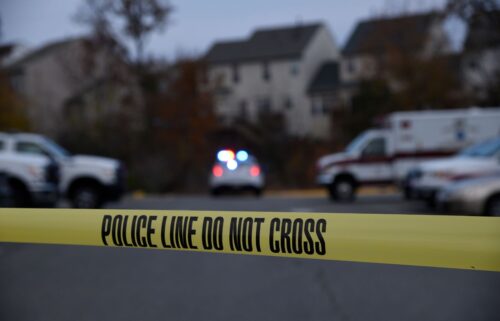 Blurred background with an ambulance, police vehicle with flashing lights, and two houses. Foreground shows yellow 