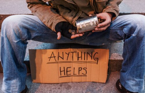 Poverty in the US. A person in worn jeans and a brown jacket sits on stairs, holding a tin can for coins. A cardboard sign reads 