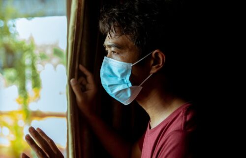 A person wearing a COVID medical mask stands by a window, gazing outside with a hand resting on the curtain. The scene is softly lit.
