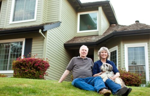 A smiling boomer couple sits on grass in front of a two-story house, with a small dog on their lap. The home is framed by windows and shrubs, creating a warm, welcoming scene.