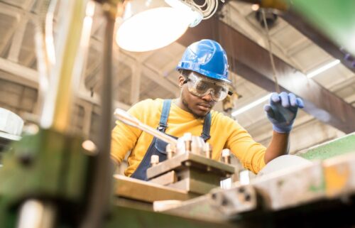 A focused worker wearing a blue hard hat and goggles operates machinery in a manufacturing workshop. Bright overhead light illuminates the industrial setting.
