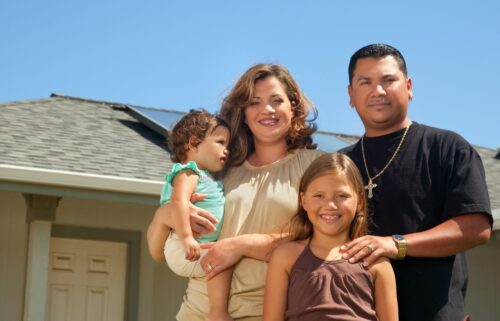A Hispanic family of four stands in front of a house with solar panels. The mother holds a young child, while the father and older daughter smile warmly.