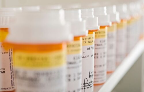 Are drug prices going to get lower? Rows of orange prescription bottles with white caps and yellow labels are organized on a shelf.