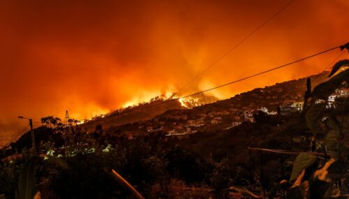 A large wildfire illuminates the night sky with orange flames on a hillside above a town. Smoke fills the air, evoking a sense of danger, disaster, and urgency. Photo by Michael Held on Unsplash