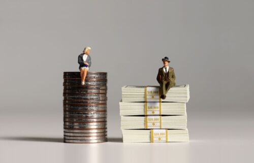 Miniature figures on stacks; a woman sits on coins, a man on paper money, symbolizing financial and pay inequality. Neutral background.