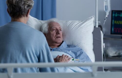 Elderly man in hospital bed, looking concerned, holds hands with a visitor seated beside him. An IV drip and heart monitor are visible, suggesting a hospital setting.