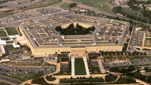 Aerial view of the Pentagon, a large, five-sided concrete building with a central courtyard. Surrounding are roads, parking lots, and green spaces.