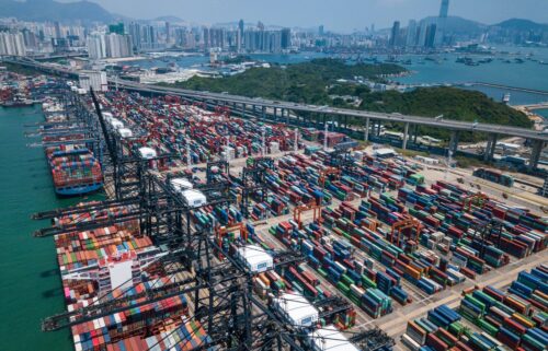 Aerial view of a bustling port with colorful shipping containers neatly arranged, representing the GDP report. Cranes line the docks with city skyscrapers and mountains in the distance