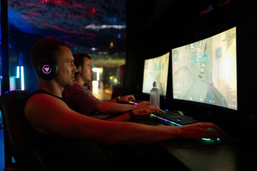 Two gamers intensely focus on computer screens in a dark room, wearing headsets. The colorful lighting and monitors create an immersive, competitive atmosphere. https://unsplash.com/photos/two-men-playing-video-games-on-computers-rtXP3mG8DBs?utm_source=unsplash&utm_medium=referral&utm_content=creditShareLink